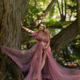 FotoArtist - Kobieta w zwiewnej, różowej sukni ciążowej pozuje na tle leśnego krajobrazu, opierając się o korę drzewa.