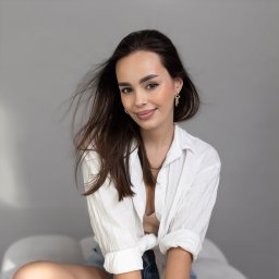 FotoArtist - Portret uśmiechniętej brunetki w białej koszuli, z delikatnym makijażem i złotymi kolczykami na tle szarej ściany.