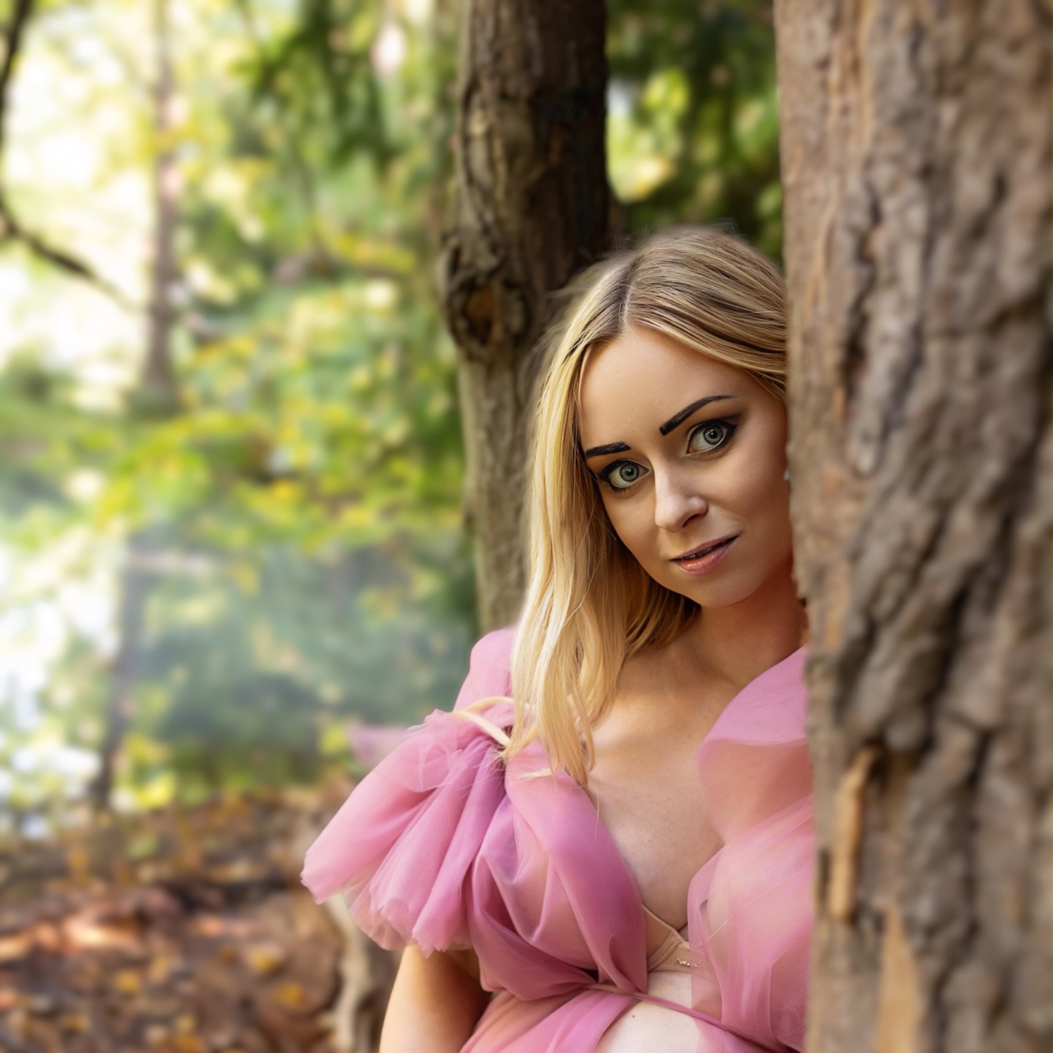 Portret kobiety w różowej, tiulowej sukni, ukrywającej się za drzewem w lesie, z wyraźnym makijażem oczu i blond włosami.