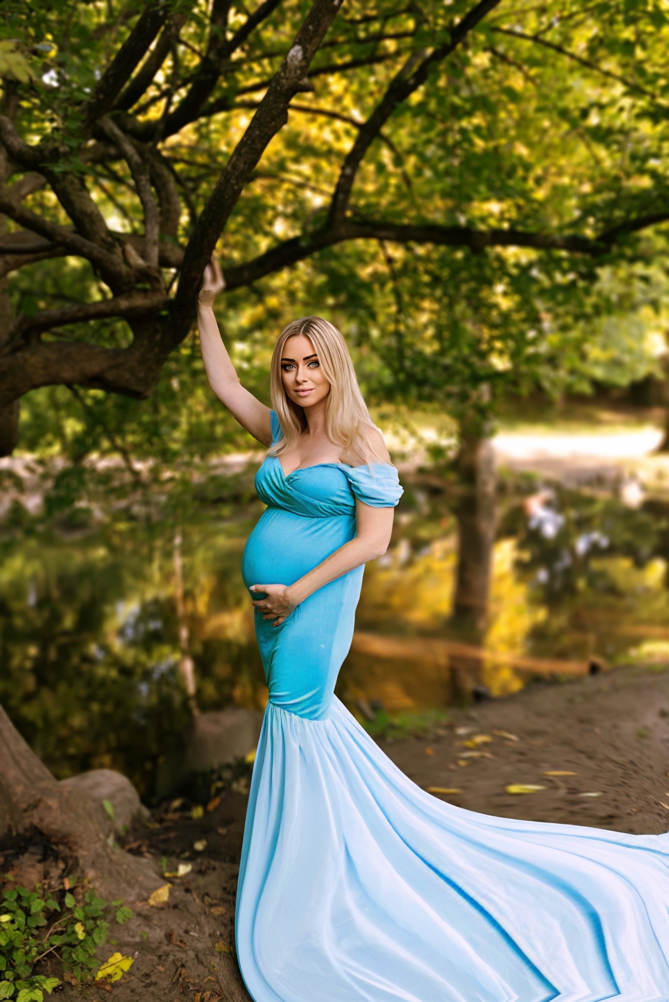 Kobieta w ciąży w błękitnej sukni z trenem pozuje na tle drzew i wody, trzymając ręce na brzuchu.