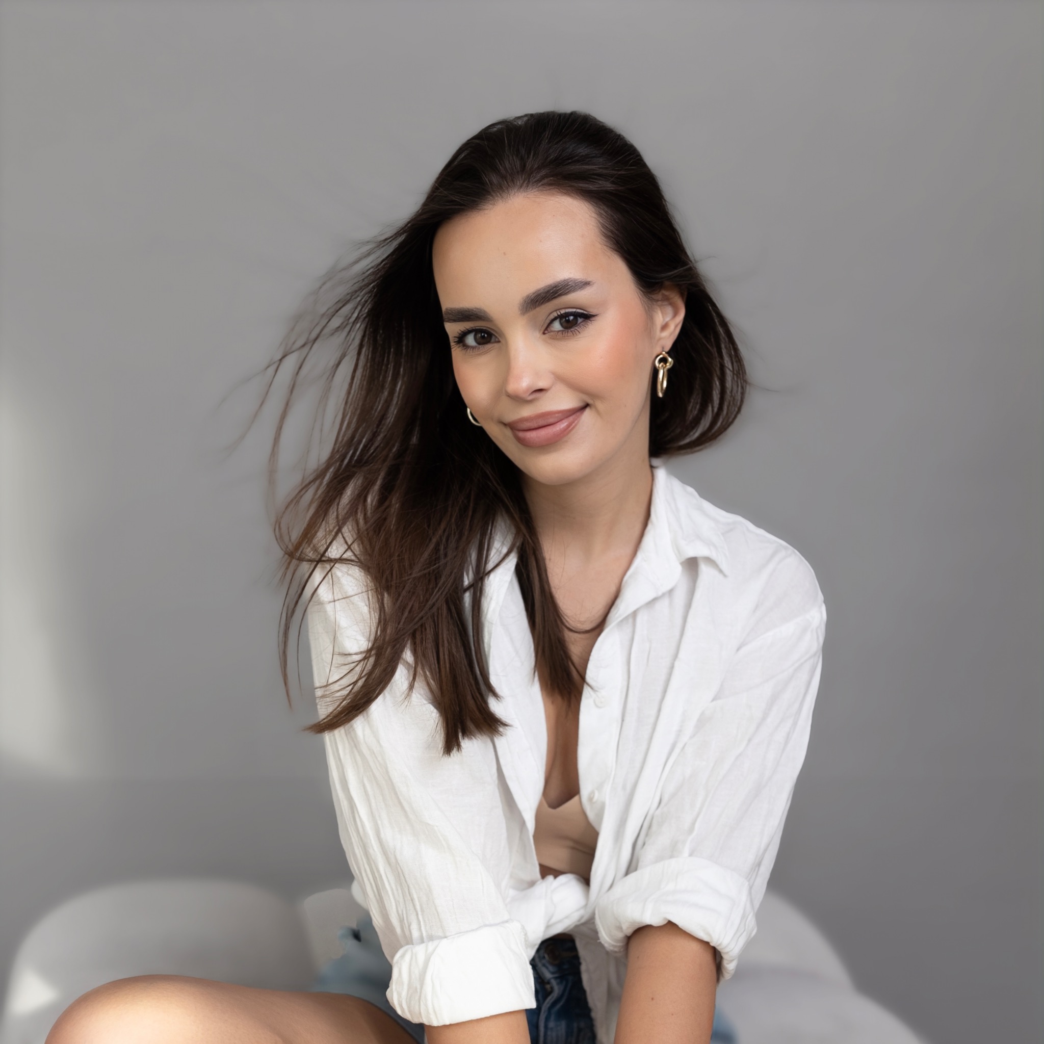 Portret uśmiechniętej brunetki w białej koszuli, z delikatnym makijażem i złotymi kolczykami na tle szarej ściany.