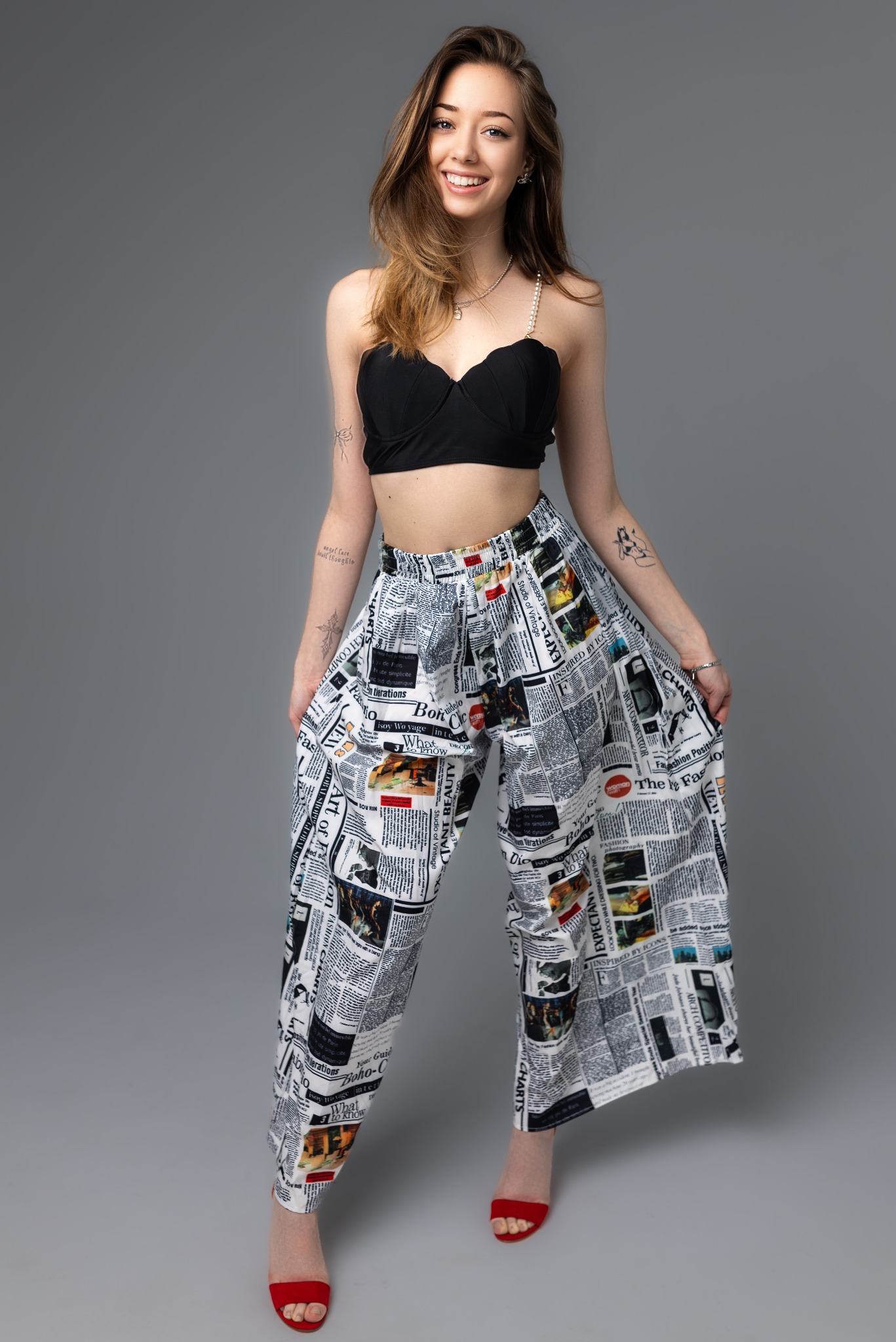 Uśmiechnięta młoda kobieta w czarnym topie i szerokich spodniach z nadrukiem gazetowym, pozująca na szarym tle. Widoczne tatuaże na ramionach i czerwone sandały na obcasie.