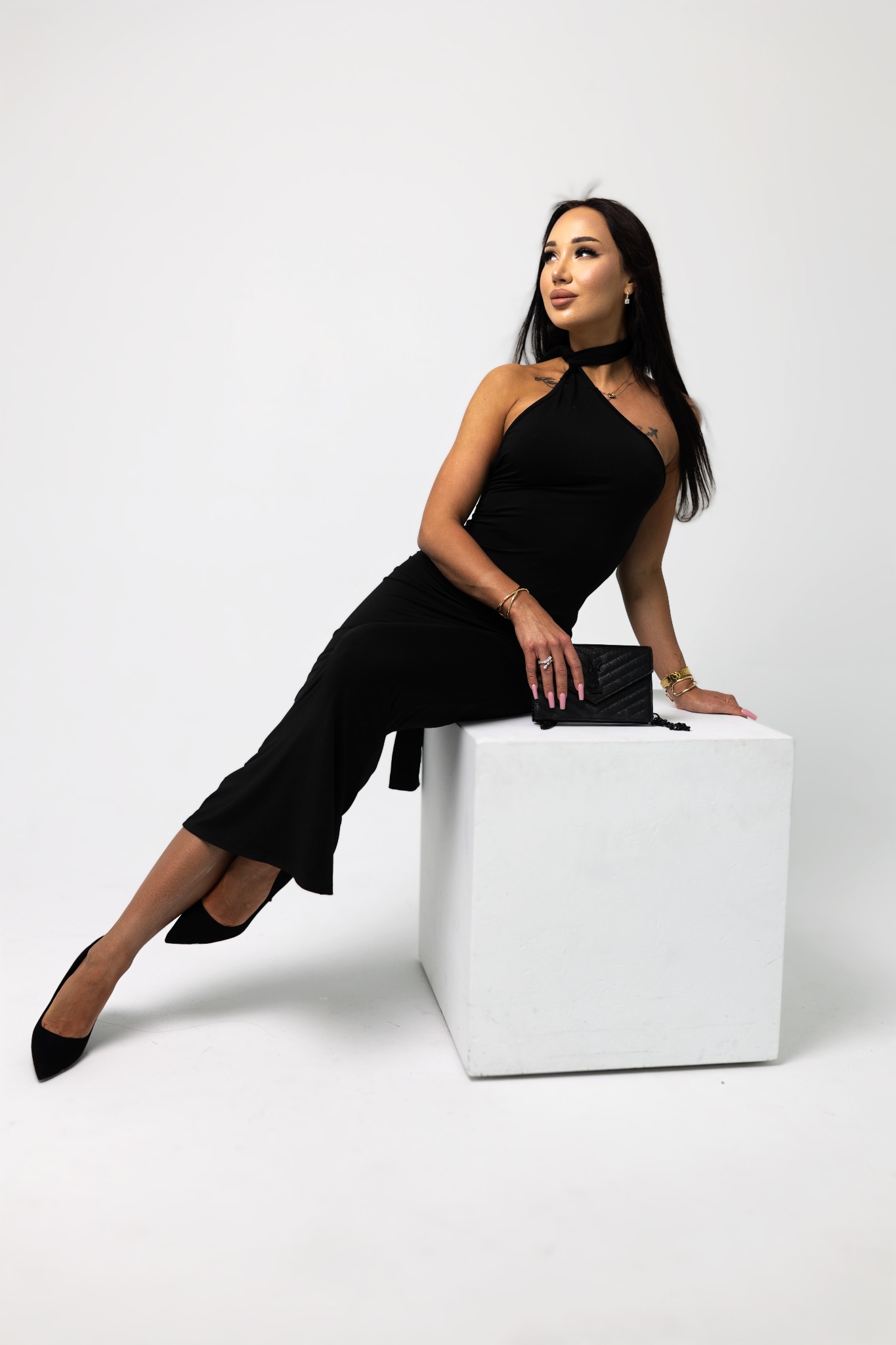 Kobieta w czarnej sukience i szpilkach pozuje siedząc na białym sześcianie w studiu w Gdańsku. Uwagę zwracają złote bransoletki i mała czarna torebka.