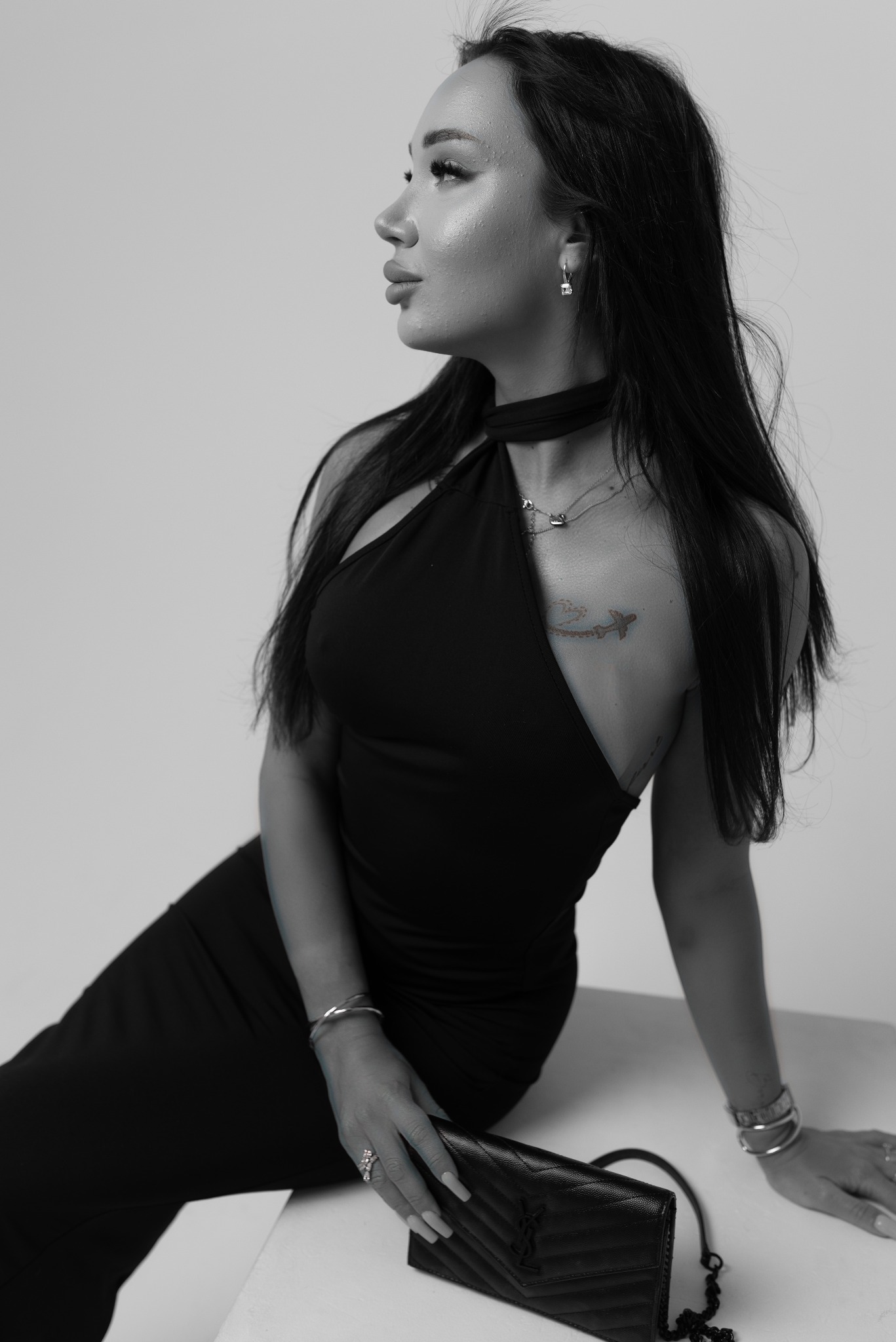 Czarno-białe zdjęcie kobiety w czarnej sukience z odkrytymi ramionami, z makijażem i biżuterią, siedzącej obok czarnej torebki na białym tle.