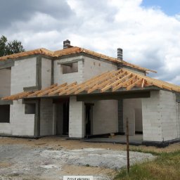 M2CAPITAL SPÓŁKA Z OGRANICZONĄ ODPOWIEDZIALNOŚCIĄ - Surowy stan domu murowanego z bloczków, widoczne drewniane elementy konstrukcji dachu, dwa kominy, tablica informacyjna budowy w prawym dolnym rogu.