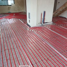 M2CAPITAL SPÓŁKA Z OGRANICZONĄ ODPOWIEDZIALNOŚCIĄ - Instalacja ogrzewania podłogowego z czerwonych rurek na folii refleksyjnej, widoczne fragmenty ścian w stanie surowym i schody drewniane.