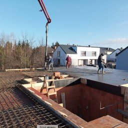 M2CAPITAL SPÓŁKA Z OGRANICZONĄ ODPOWIEDZIALNOŚCIĄ - Wylewanie betonu na strop domu jednorodzinnego z widocznym zbrojeniem i deskowaniem, kilku robotników w trakcie pracy, w tle zabudowa mieszkalna i las.