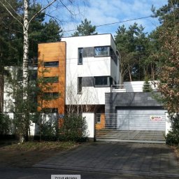 M2CAPITAL SPÓŁKA Z OGRANICZONĄ ODPOWIEDZIALNOŚCIĄ - Nowoczesny dom z elewacją w bieli i drewnie, z garażem wykończonym szarym kamieniem, otoczony zielenią i ogrodzeniem z metalowymi przęsłami.