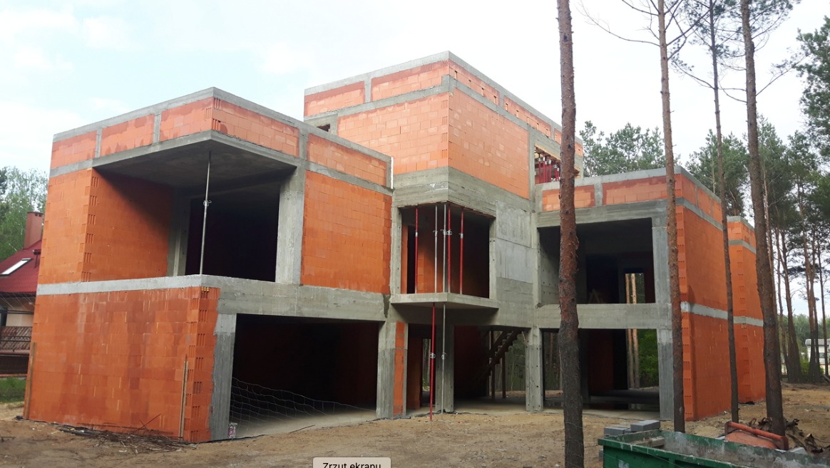 Surowy stan domu murowanego z betonu i cegły ceramicznej, widoczne podpory stropu i schody zewnętrzne, otoczenie w stanie budowy, wysokie drzewa w tle.