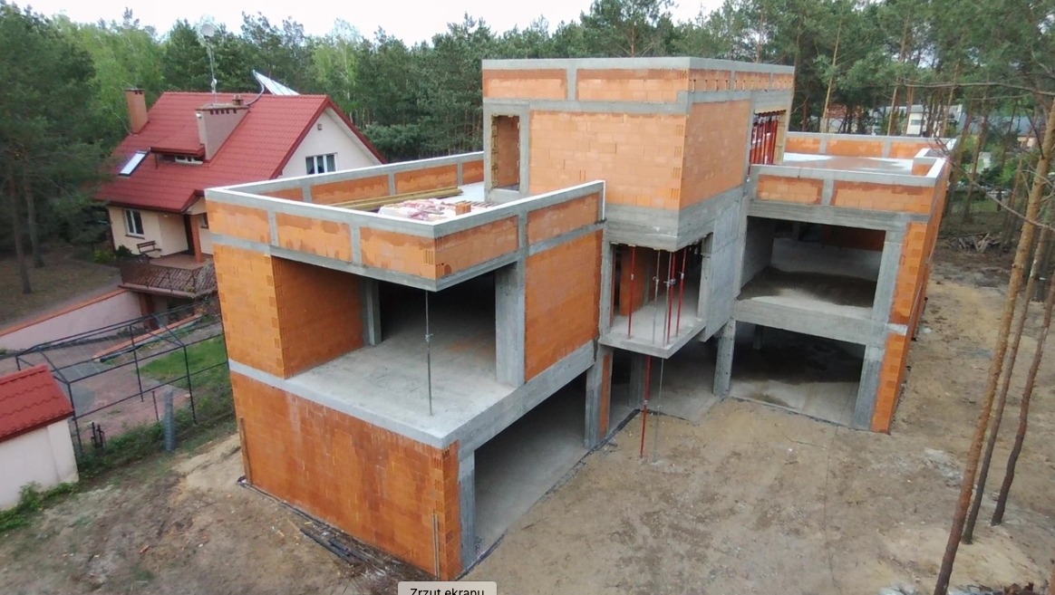 Surowy, dwupiętrowy dom z czerwonej cegły i betonu w trakcie budowy, widok z góry, z sąsiednim domem z czerwonym dachem w tle i lasem w oddali.