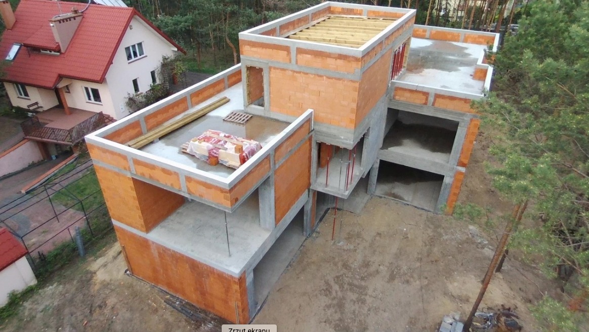 Surowy stan dwukondygnacyjnego domu murowanego z betonu i czerwonej cegły, widok z góry na plac budowy z widocznym drewnianym stropem, stertą materiałów budowlanych pod folią i sąsiednim domem.