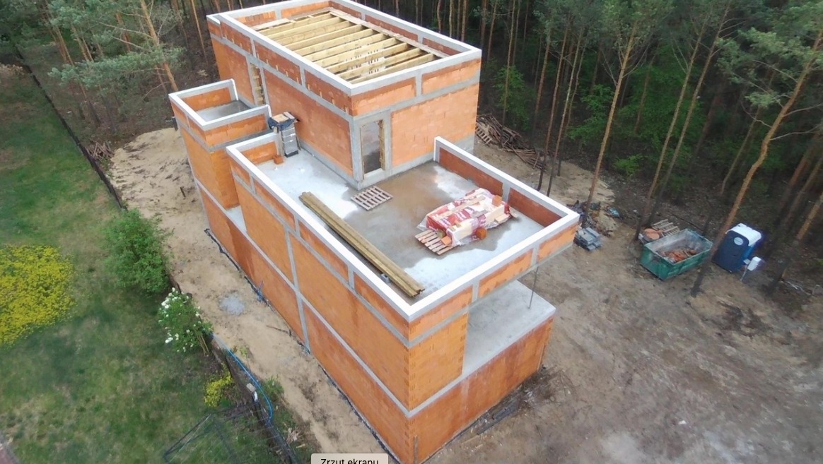 Surowy, dwukondygnacyjny dom z czerwonej cegły w trakcie budowy, widok z góry, z widocznymi materiałami budowlanymi na betonowym dachu, drewnianą konstrukcją stropu i kontenerem na gruz w tle.
