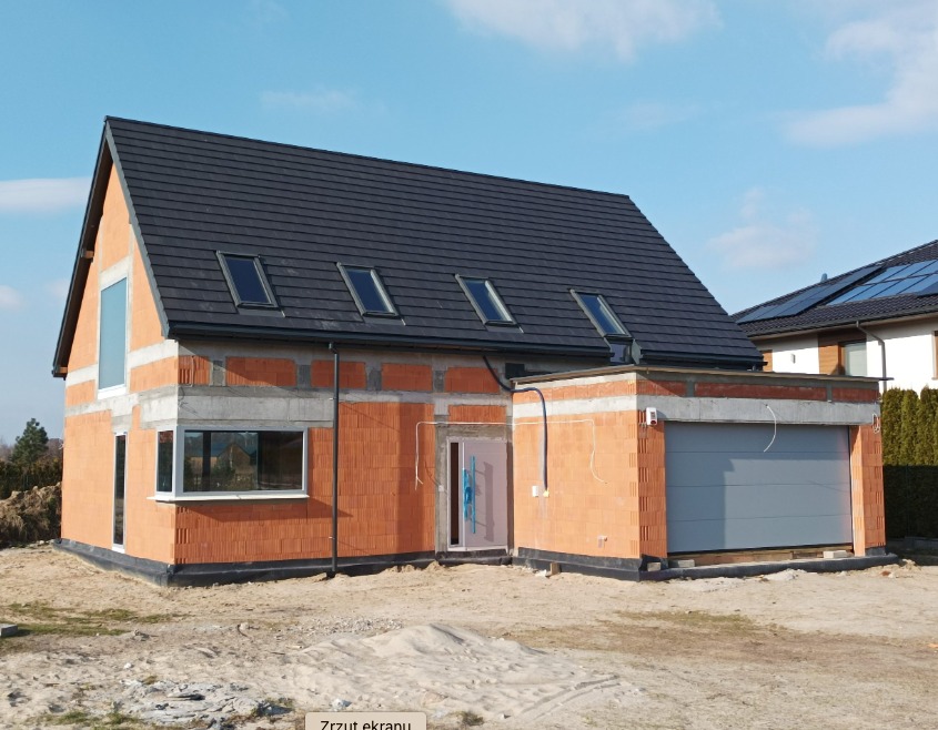 Dom jednorodzinny w trakcie budowy, widoczne ściany z czerwonej cegły, czarny dach z oknami dachowymi, brama garażowa i okna bez wykończenia, na zewnątrz piasek.
