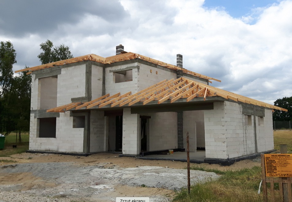 Surowy stan domu murowanego z bloczków, widoczne drewniane elementy konstrukcji dachu, dwa kominy, tablica informacyjna budowy w prawym dolnym rogu.