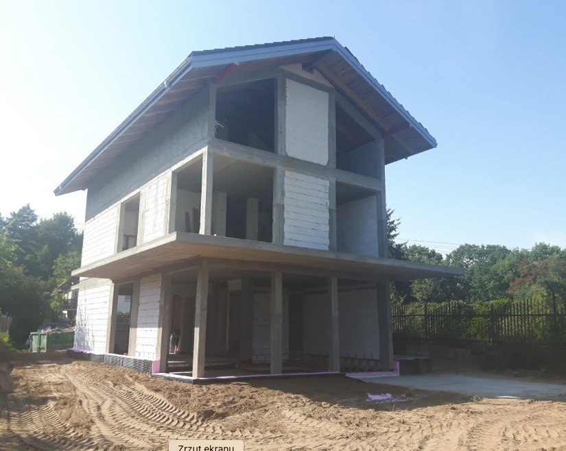 Dwukondygnacyjny dom w stanie surowym otwartym, widoczne białe bloczki, betonowy strop i więźba dachowa, teren wokół z rozjeżdżoną ziemią.