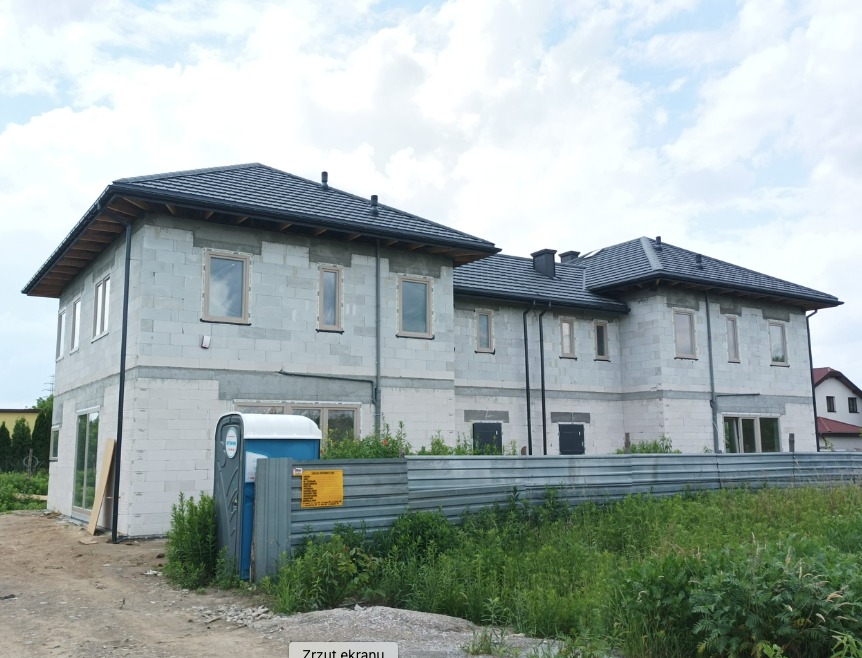 Dwukondygnacyjny dom murowany w stanie surowym zamkniętym z grafitową dachówką, widoczne okna, szara elewacja z betonu komórkowego, ogrodzenie tymczasowe i toaleta przenośna.
