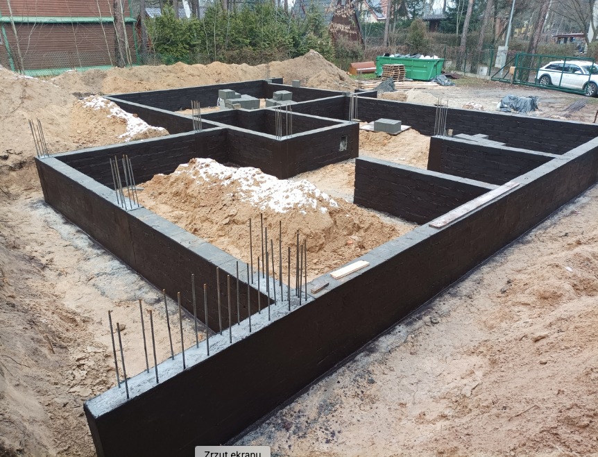 Wylane fundamenty domu z wystającymi prętami zbrojeniowymi, zabezpieczone czarną izolacją bitumiczną, częściowo zasypane piaskiem.