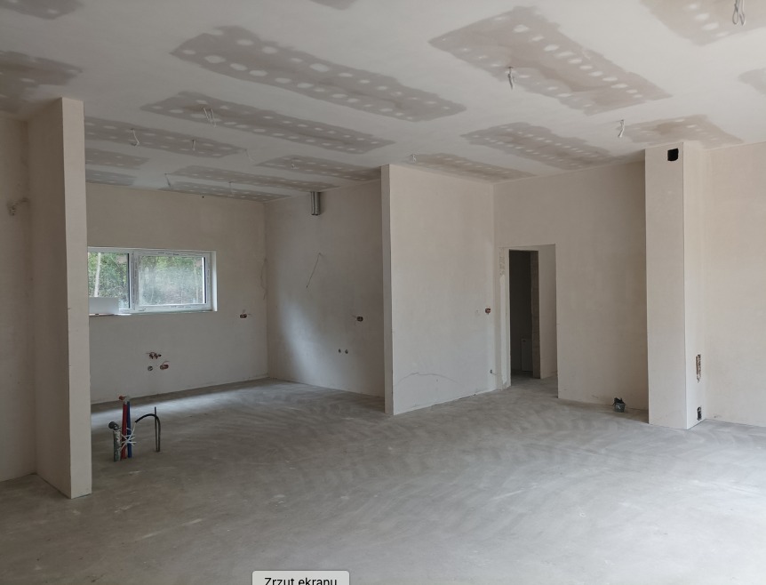 Wnętrze w stanie deweloperskim z surową podłogą, ścianami przygotowanymi do malowania i sufitem z widocznymi śladami po szpachlowaniu płyt gipsowo-kartonowych, instalacja hydrauliczna wystaje ze...