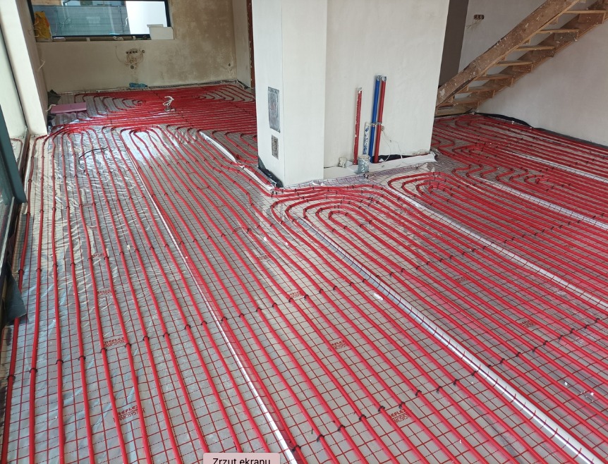 Instalacja ogrzewania podłogowego z czerwonych rurek na folii refleksyjnej, widoczne fragmenty ścian w stanie surowym i schody drewniane.