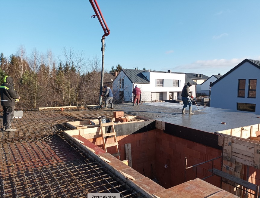 Wylewanie betonu na strop domu jednorodzinnego z widocznym zbrojeniem i deskowaniem, kilku robotników w trakcie pracy, w tle zabudowa mieszkalna i las.