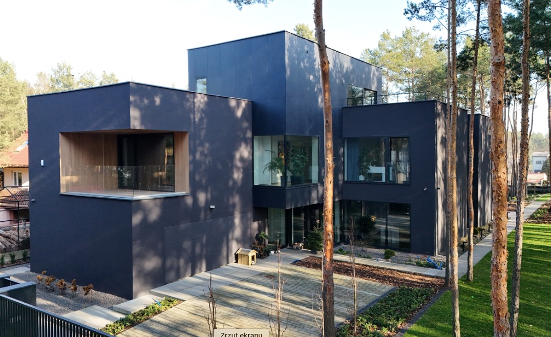 Nowoczesny dom w ciemnej kolorystyce z płaskim dachem, dużymi oknami i balkonem, otoczony drzewami iglastymi i starannie zagospodarowanym terenem.