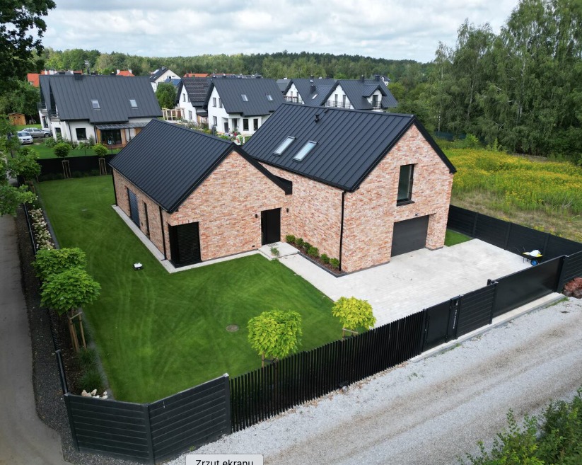Nowoczesny dom murowany z czerwoną cegłą, czarnym dachem i ogrodzeniem, widok z góry na zadbany trawnik i otoczenie.