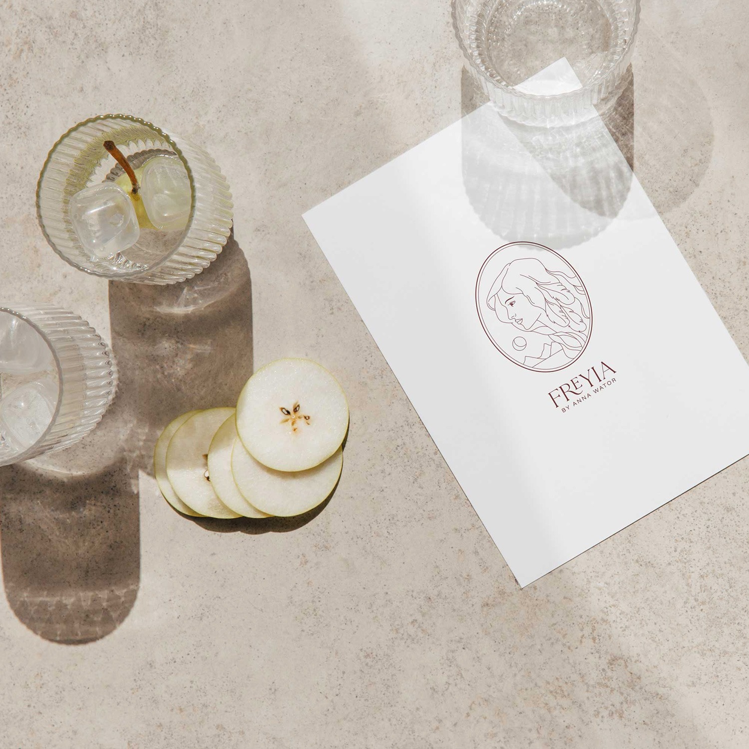 Grafika logo Freyia na białej kartce, obok szklanki z lodem i plasterków gruszki na beżowym tle w naturalnym świetle.