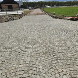 Ułożona kostka granitowa w łukowaty wz&oacute;r tworzy drogę w kierunku budynku i zielonego pola, pod błękitnym niebem z chmurami.