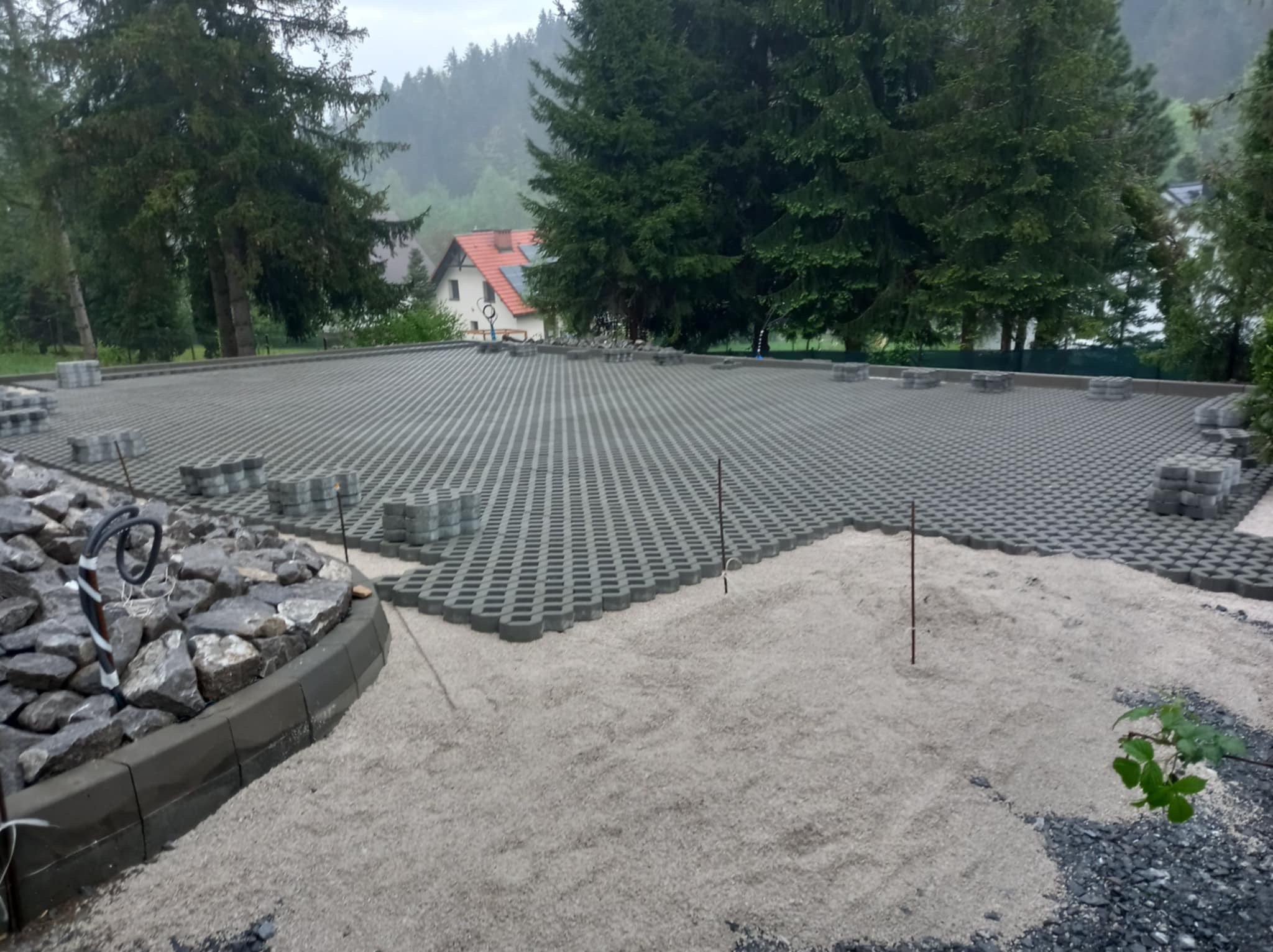 Przygotowany teren pod budowę parkingu z wykorzystaniem ażurowych płyt betonowych, częściowo zasypany piaskiem, obrzeże z kamieni i kostki betonowej, w tle zalesione wzgórze i dom.