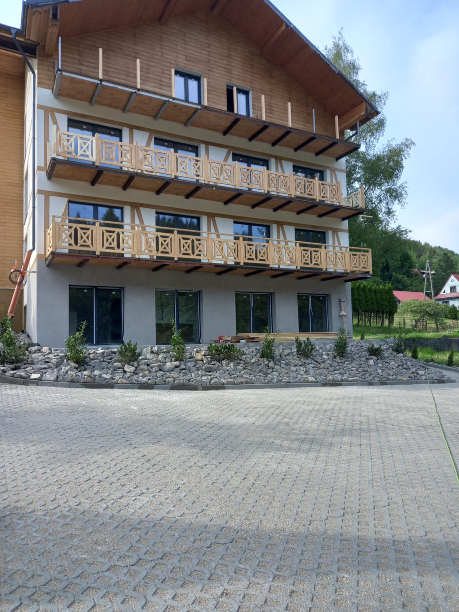 Trzypiętrowy budynek z drewnianą elewacją i balkonami z drewnianymi balustradami, otoczony szarą kostką ażurową i rabatą z kamieni i krzewów.