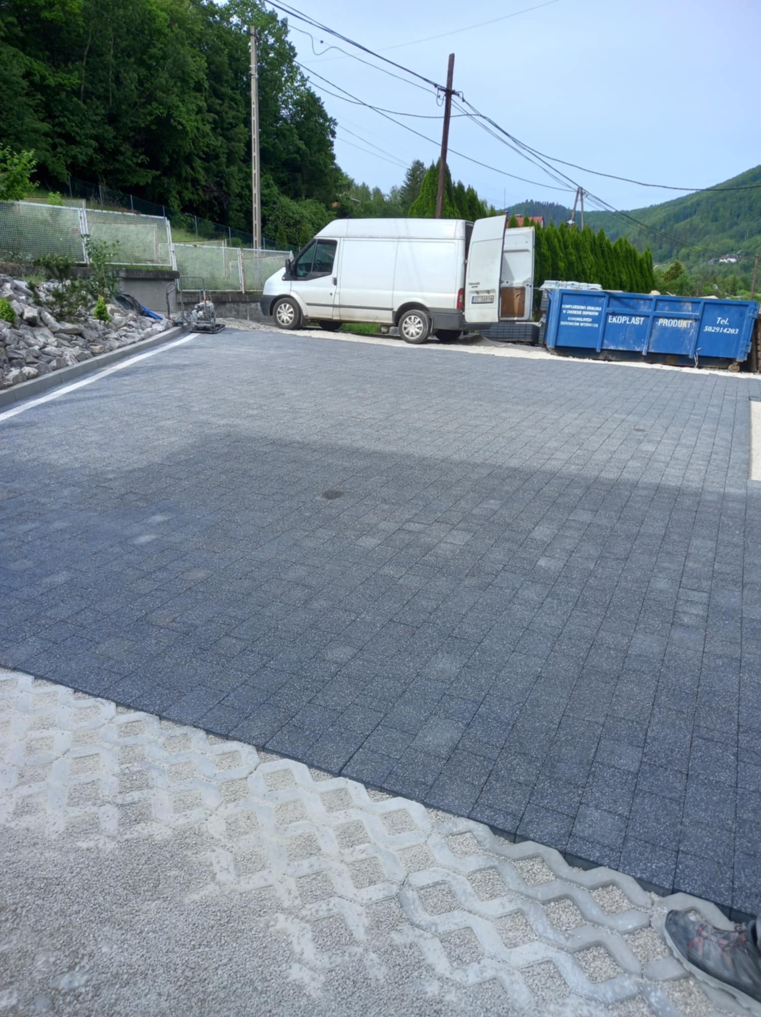Ułożona, szara kostka brukowa na posesji, z widocznymi ażurowymi płytami betonowymi po prawej stronie, biały bus i niebieski kontener w tle.