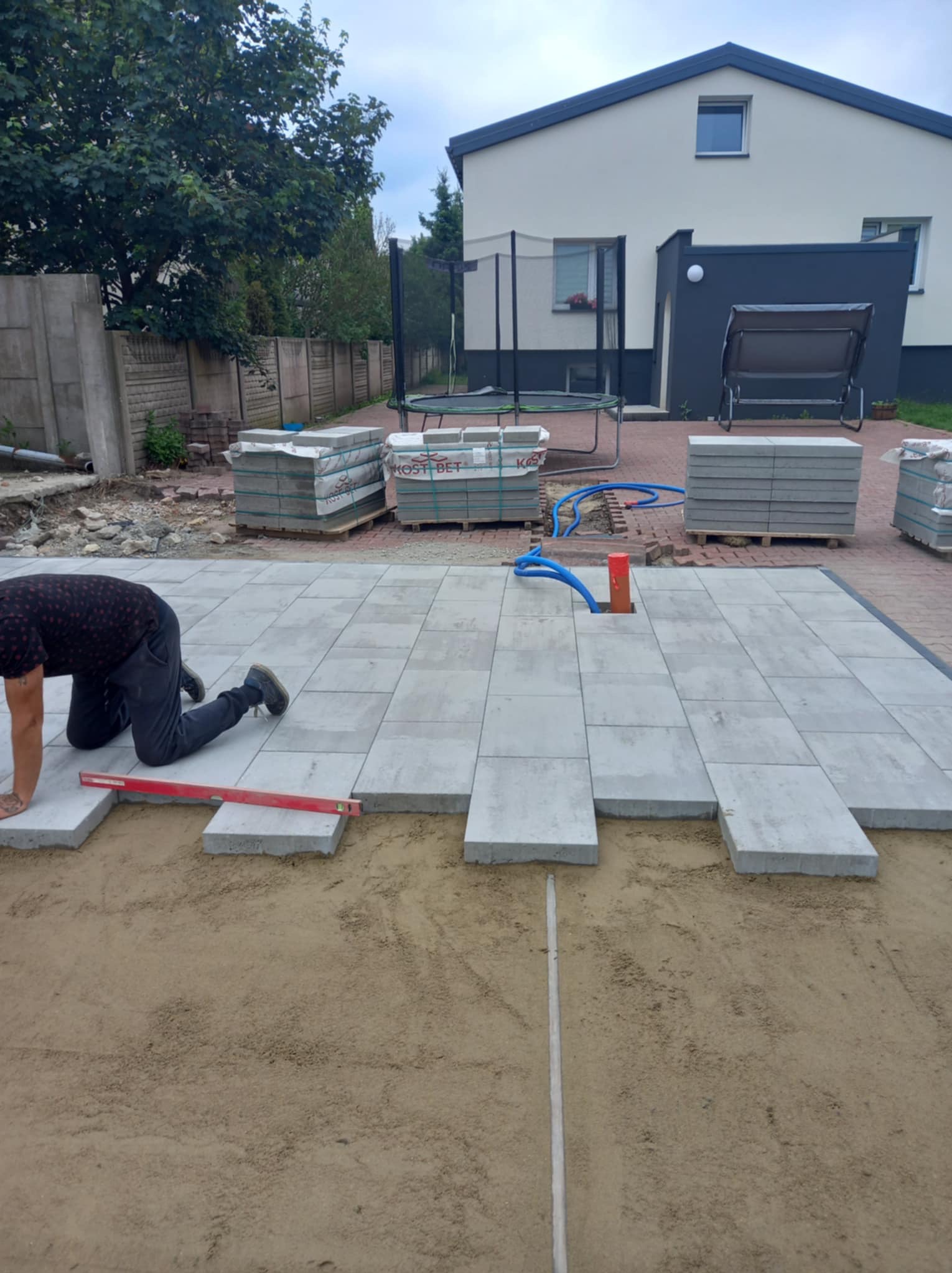 Prace brukarskie w toku: układanie szarej, prostokątnej kostki na podsypce piaskowej, widoczny poziomica i palety kostki w tle, dom z trampoliną w ogrodzie.