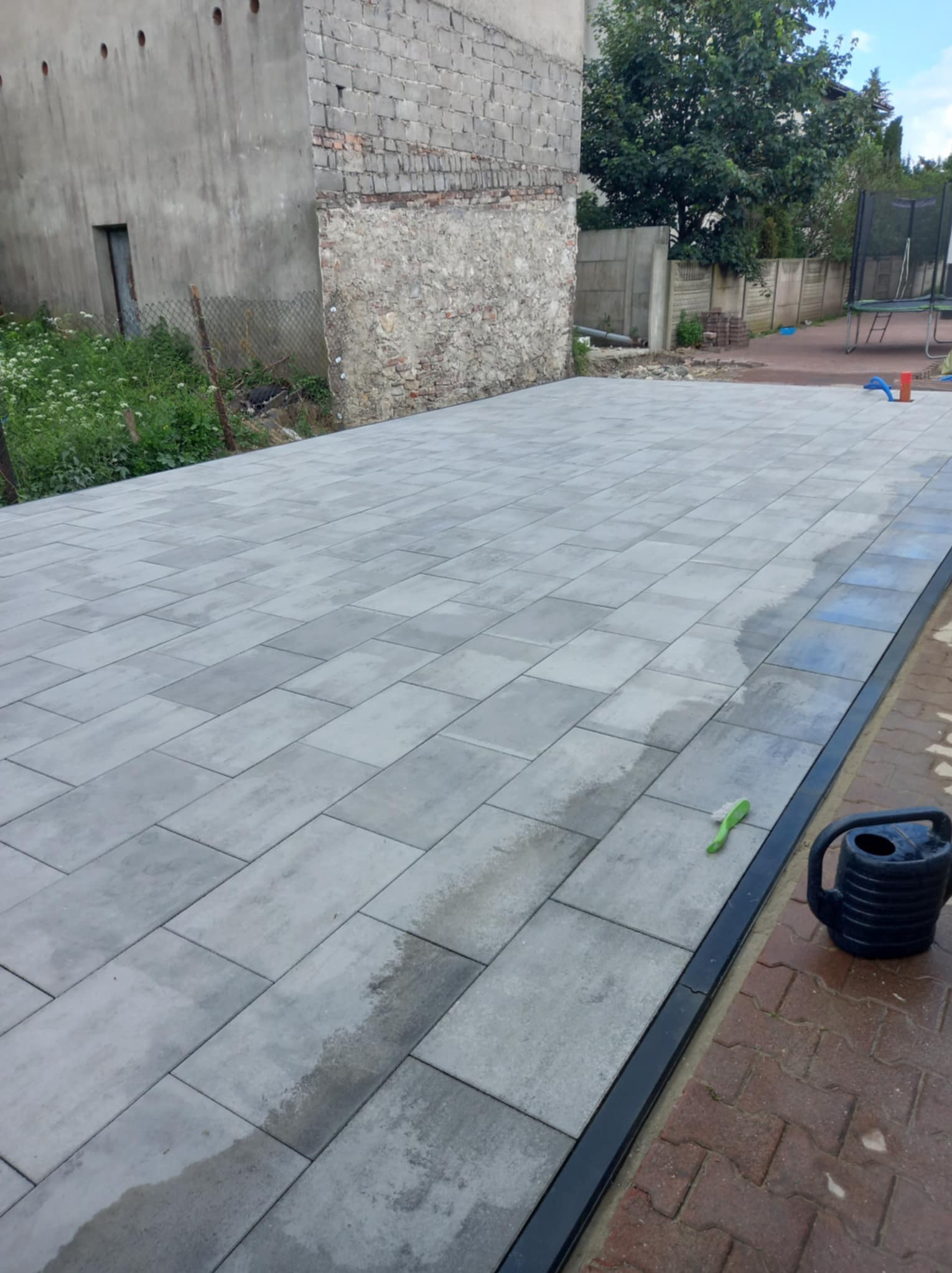 Ułożona szara kostka brukowa na posesji z widocznym starym budynkiem w tle, obok leży zielona szczotka i niebieska konewka.