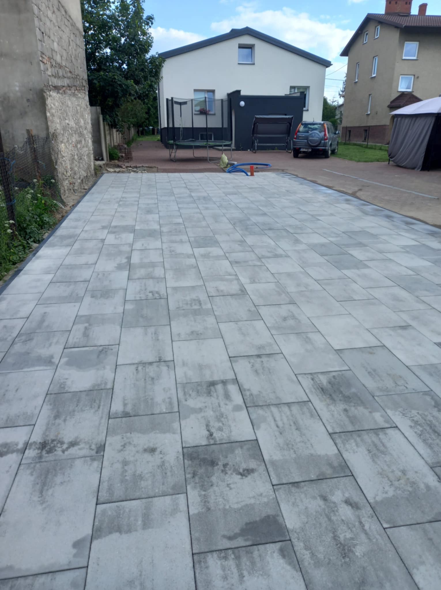 Nowo ułożona szara kostka brukowa na podwórku, widoczna granica z roślinnością i fragmenty budynków w tle.