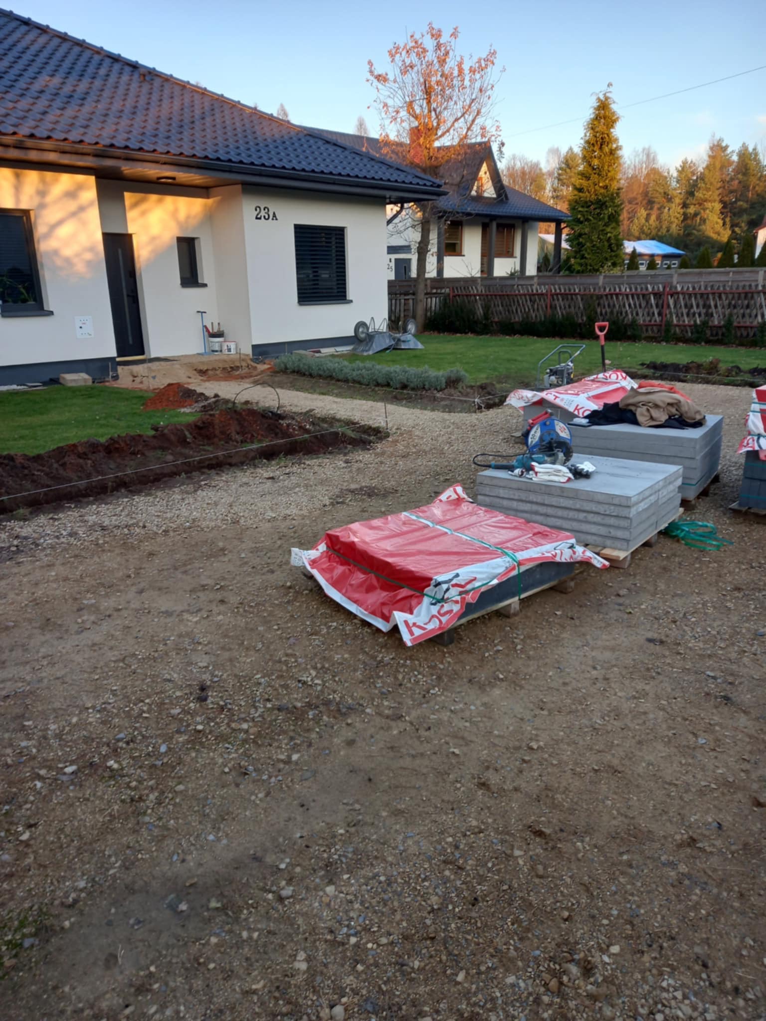 Przygotowanie terenu pod ścieżkę z kostki brukowej: wykopany rów, rozplantowany piasek, palety z kostką brukową zabezpieczone czerwoną folią, widoczna przecinarka do kostki, w tle dom z numerem 23A...