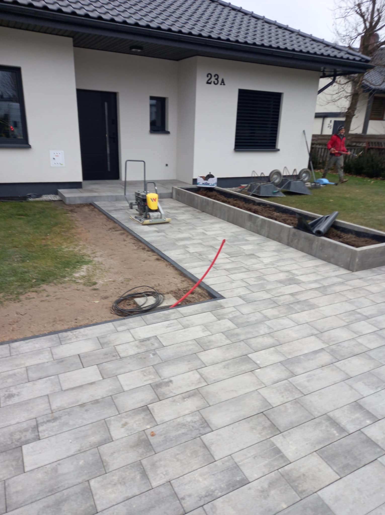 Szara kostka brukowa ułożona przed domem z numerem 23A, widoczna zagęszczarka wibracyjna, czarny kabel i fragment rabaty z ziemią, w tle osoba z narzędziem ogrodniczym.