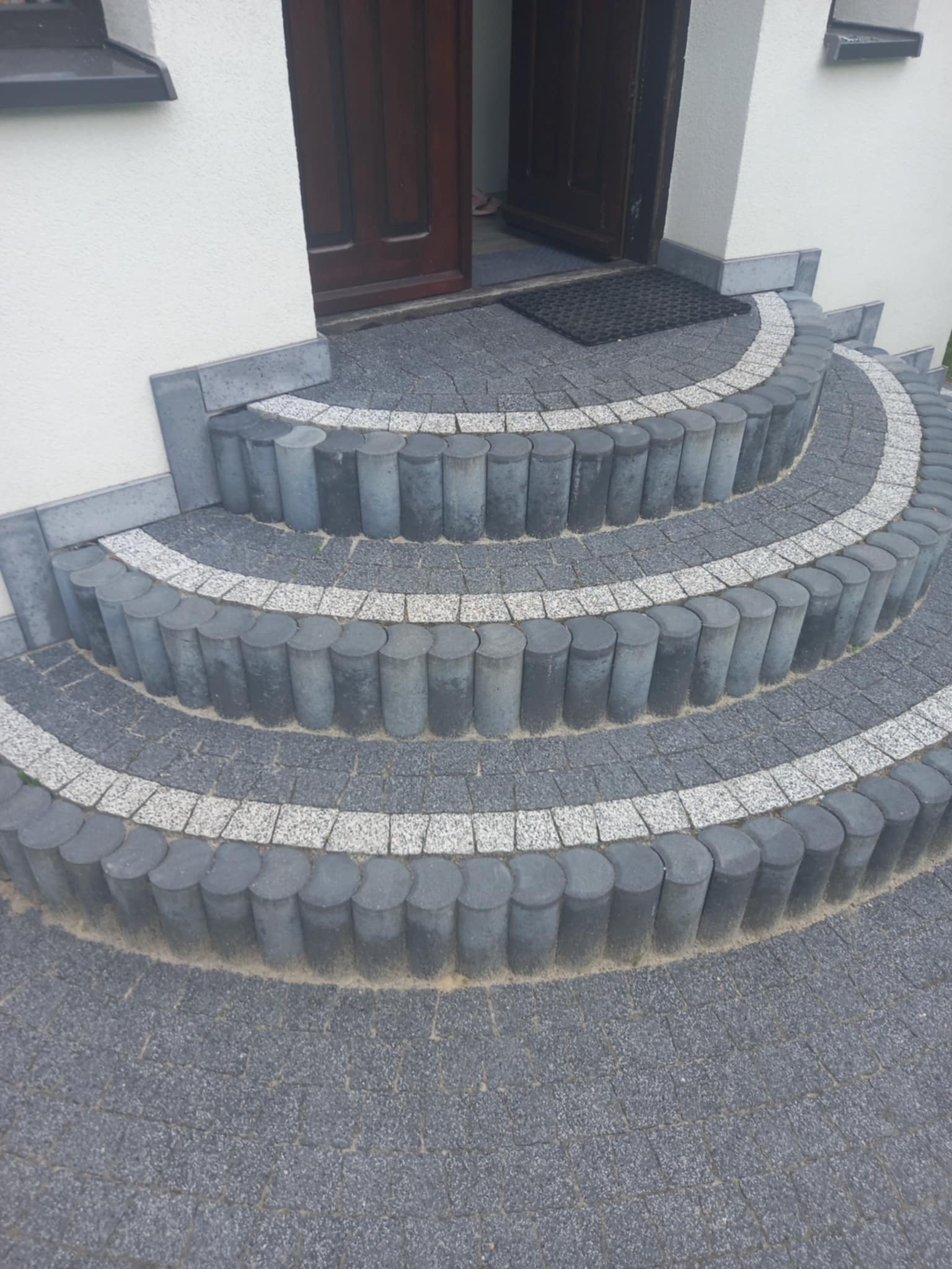 Półokrągłe schody wejściowe z kostki brukowej w odcieniach szarości, obrzeża z okrąglaków, widok z góry na drzwi wejściowe.
