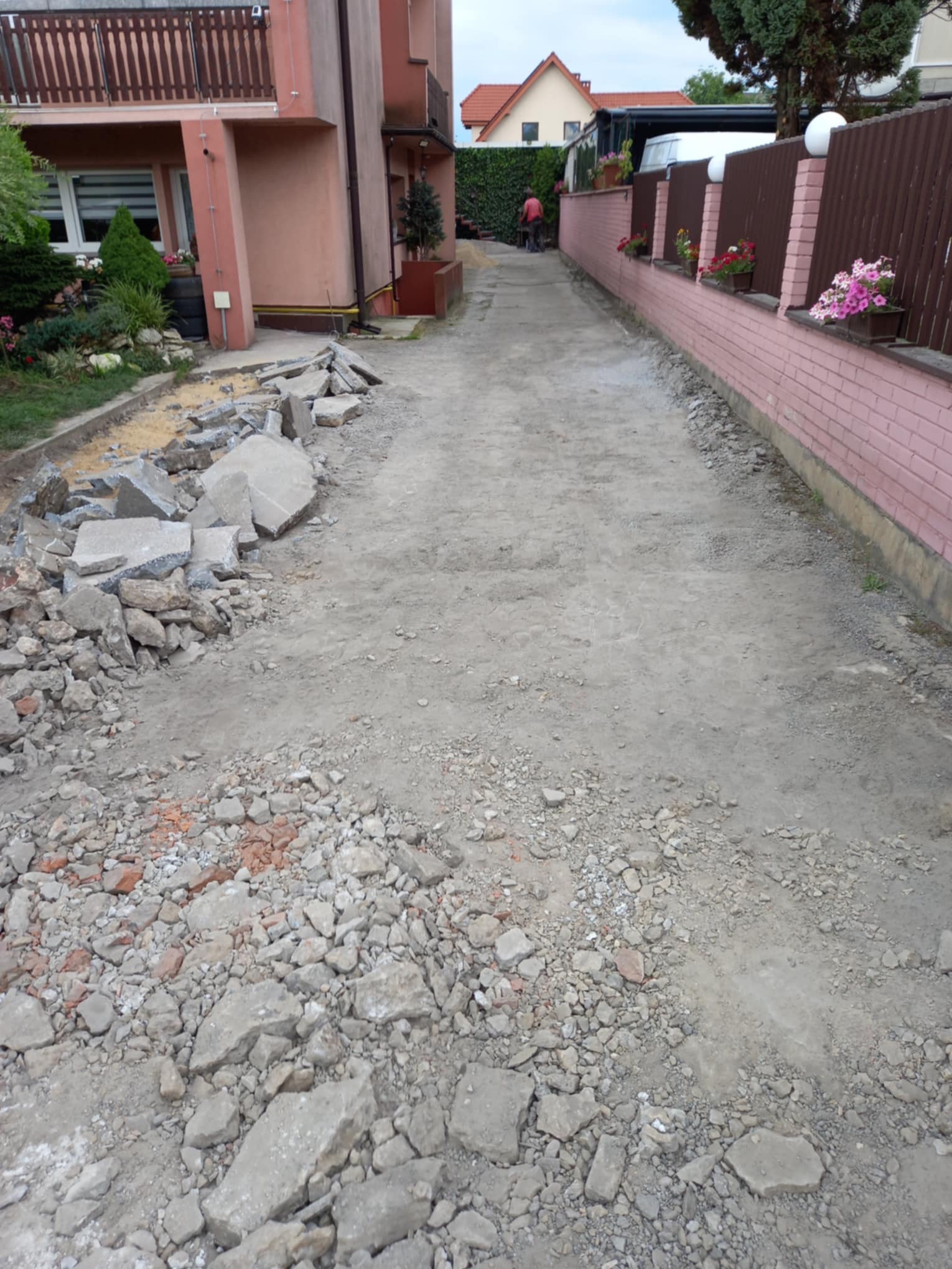 Przygotowanie podłoża pod nawierzchnię: wyrównany teren z gruzem, fragmenty zdemontowanej kostki po lewej stronie, w tle budynek mieszkalny i ogrodzenie z kwiatami.