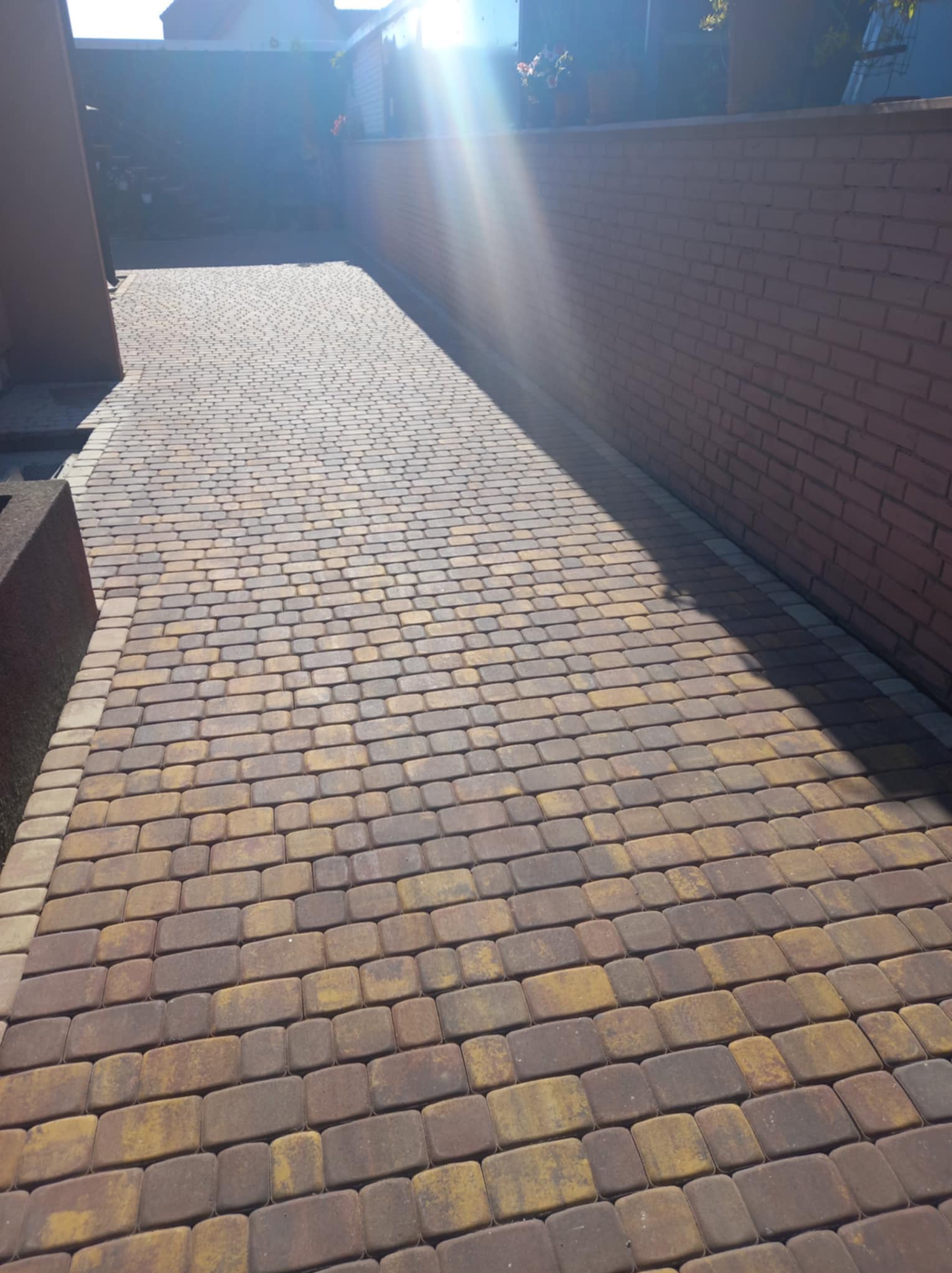 Ułożona kostka brukowa w odcieniach brązu i żółci, tworząca podjazd wzdłuż ceglanego muru, oświetlona silnym słońcem.