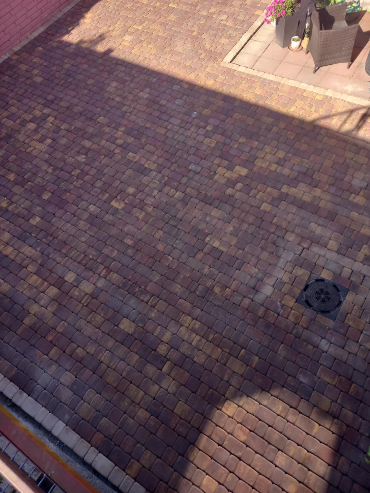 Ułożona kostka brukowa w odcieniach brązu i żółci, z widoczną studzienką kanalizacyjną i zacienionym fragmentem, otoczona fragmentem tarasu z meblami ogrodowymi i kwiatami.