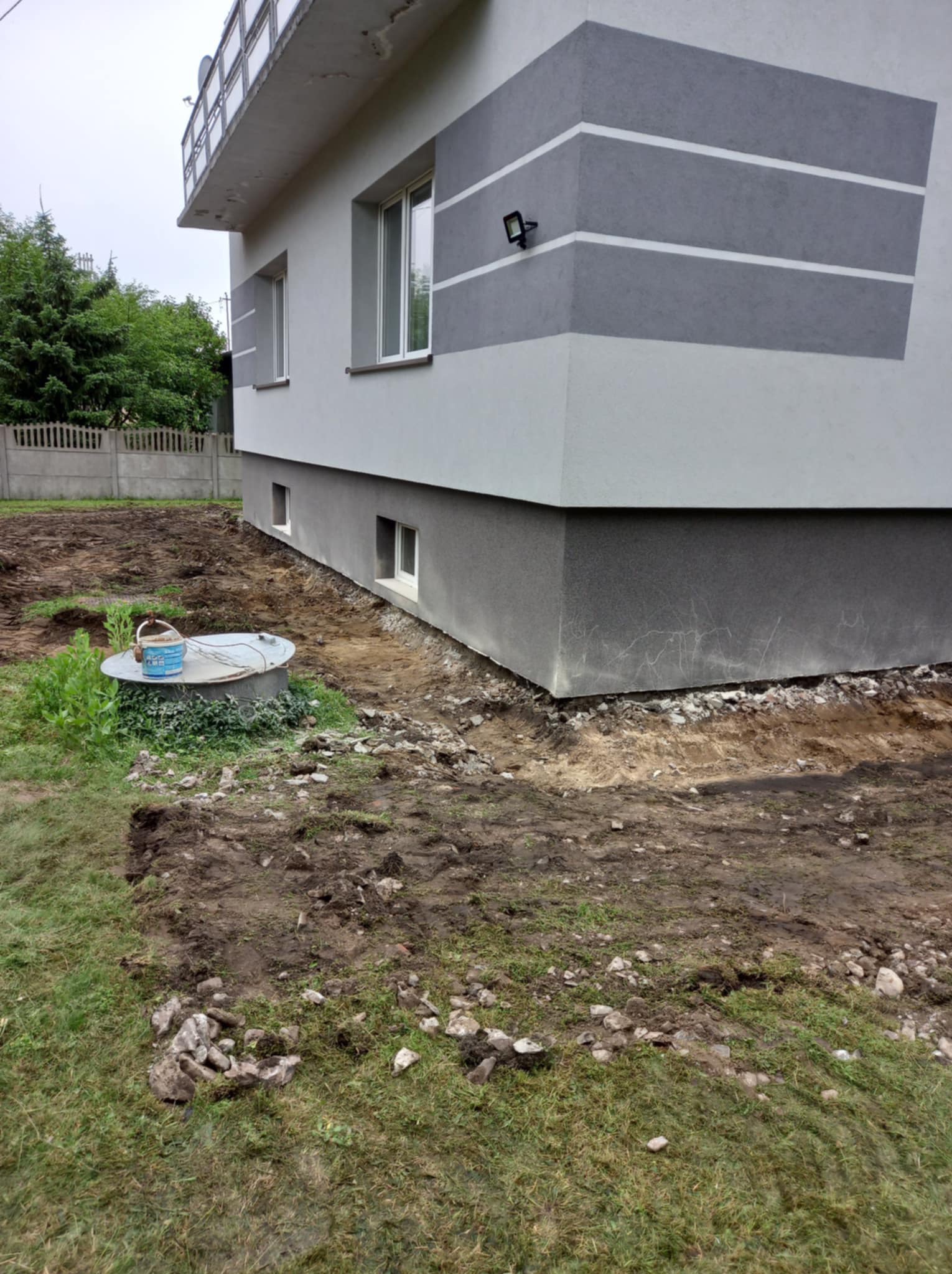 Przygotowanie terenu przy fundamencie domu: wykop pod przyszłą ścieżkę lub drenaż, widoczny fragment budynku z oknami i szarym pasem dekoracyjnym, studzienka kanalizacyjna z wiadrem farby na pokrywie.