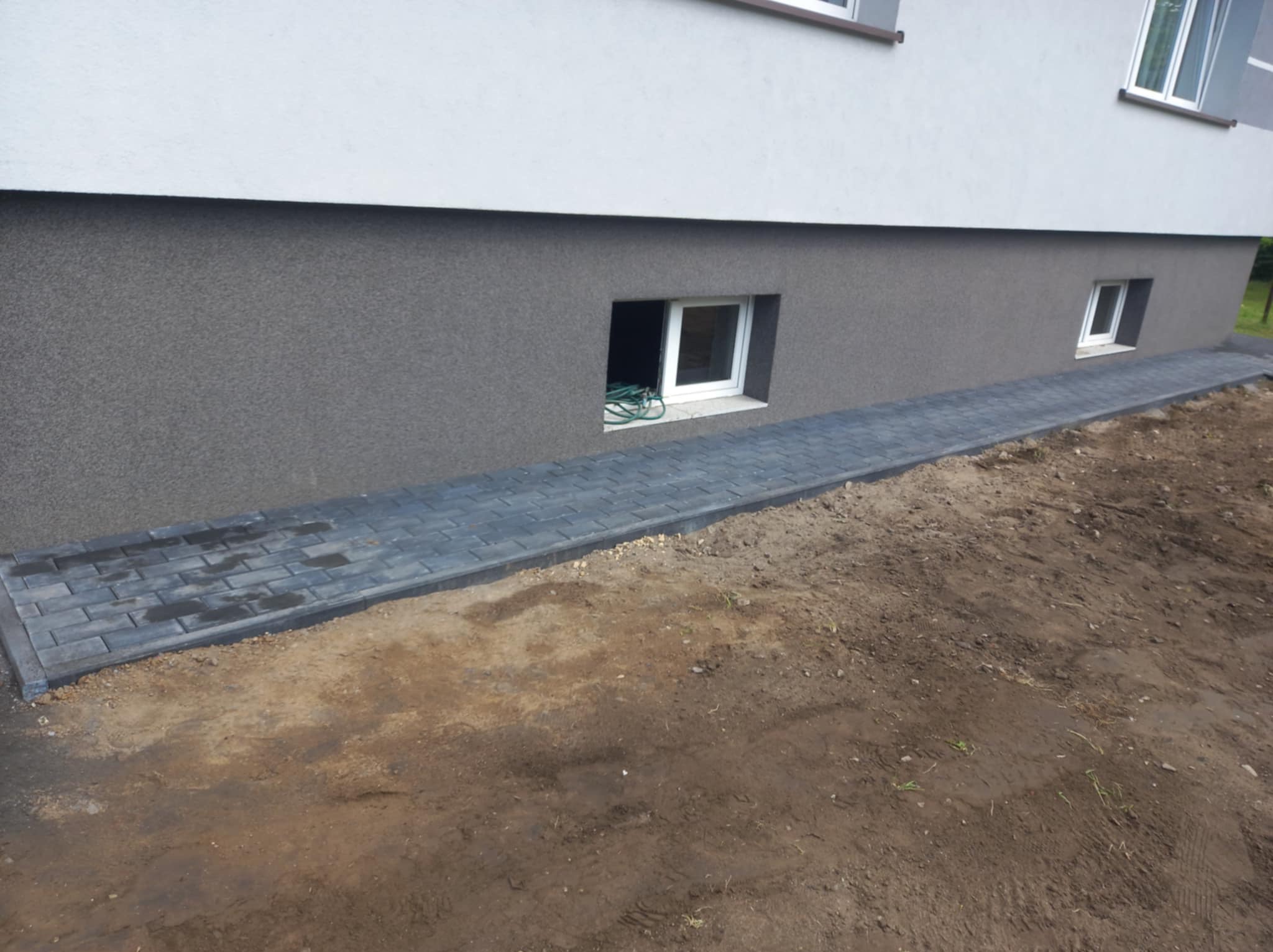 Ułożona ścieżka z szarej kostki brukowej wzdłuż fundamentu budynku z oknami piwnicznymi, widoczne ślady wilgoci na kostce i świeżo usypana ziemia po bokach.