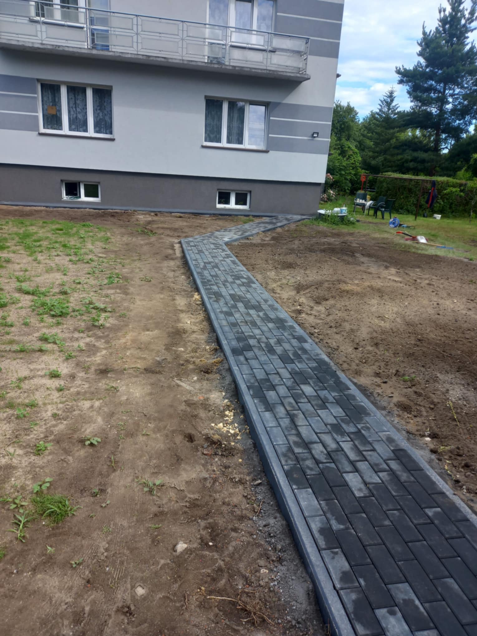 Świeżo ułożona, ciemnoszara ścieżka z kostki brukowej prowadząca od budynku w głąb ogrodu, z widocznymi śladami prac ziemnych po bokach.