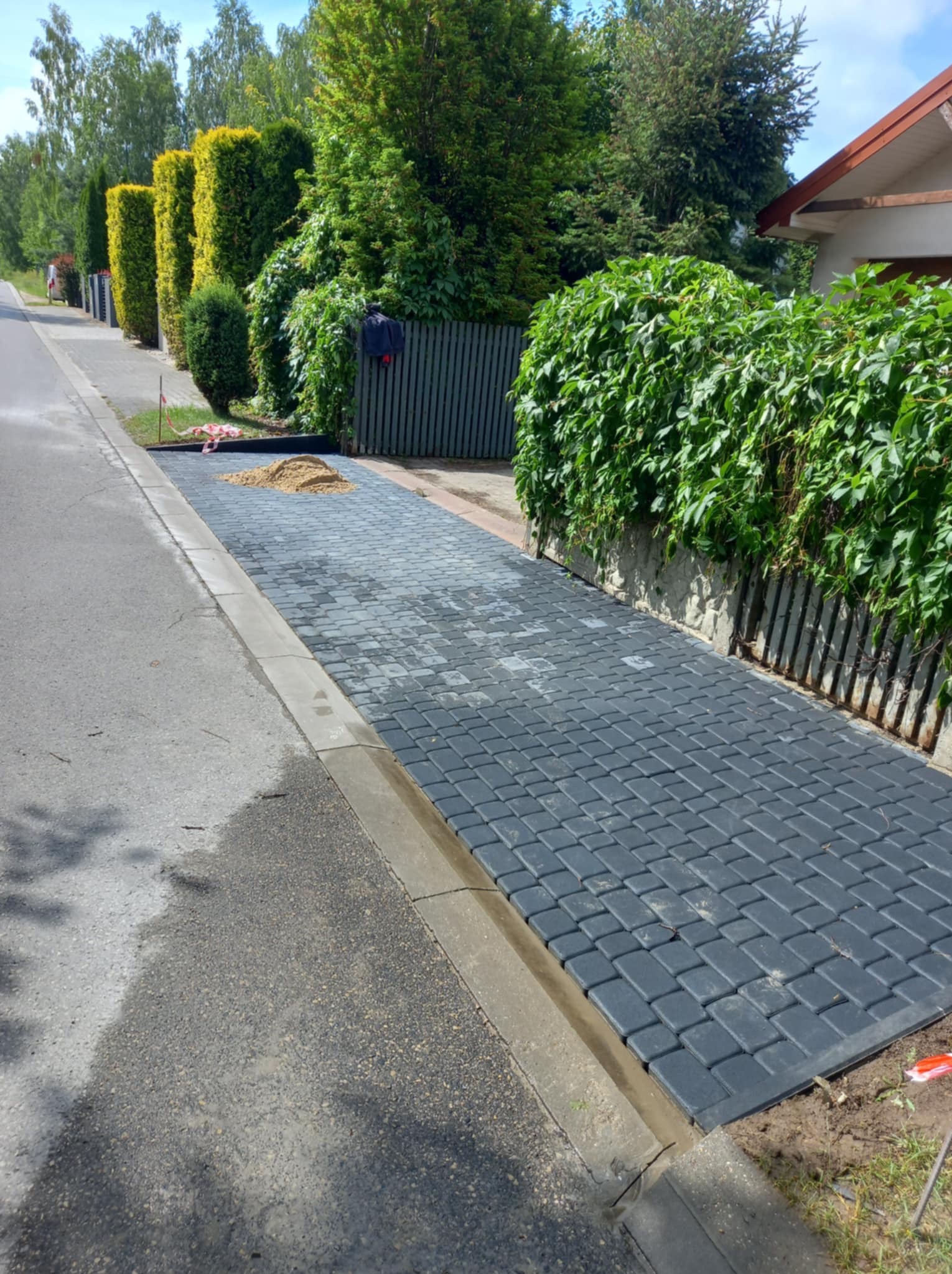 Częściowo ułożona, ciemnoszara kostka brukowa na podjeździe obok asfaltowej drogi, z widoczną stertą piasku na powierzchni i zielonym żywopłotem w tle.