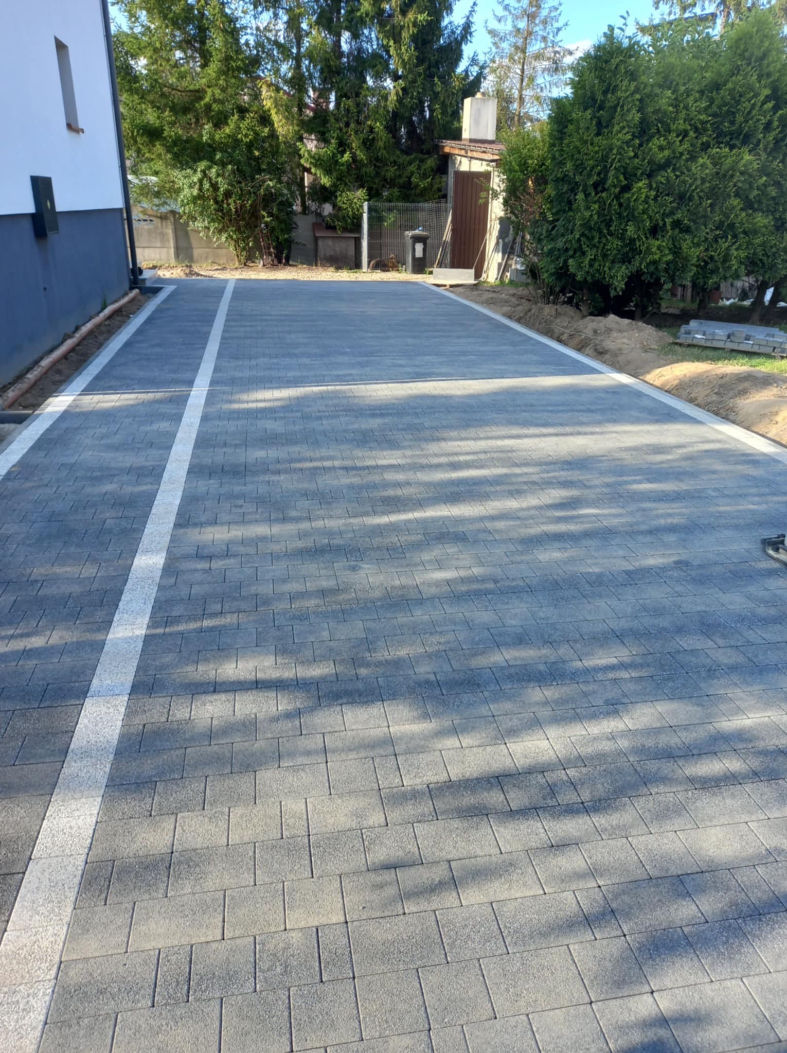 Nowo ułożony, szary podjazd z kostki brukowej z białymi pasami oddzielającymi, widoczne drzewa w tle.