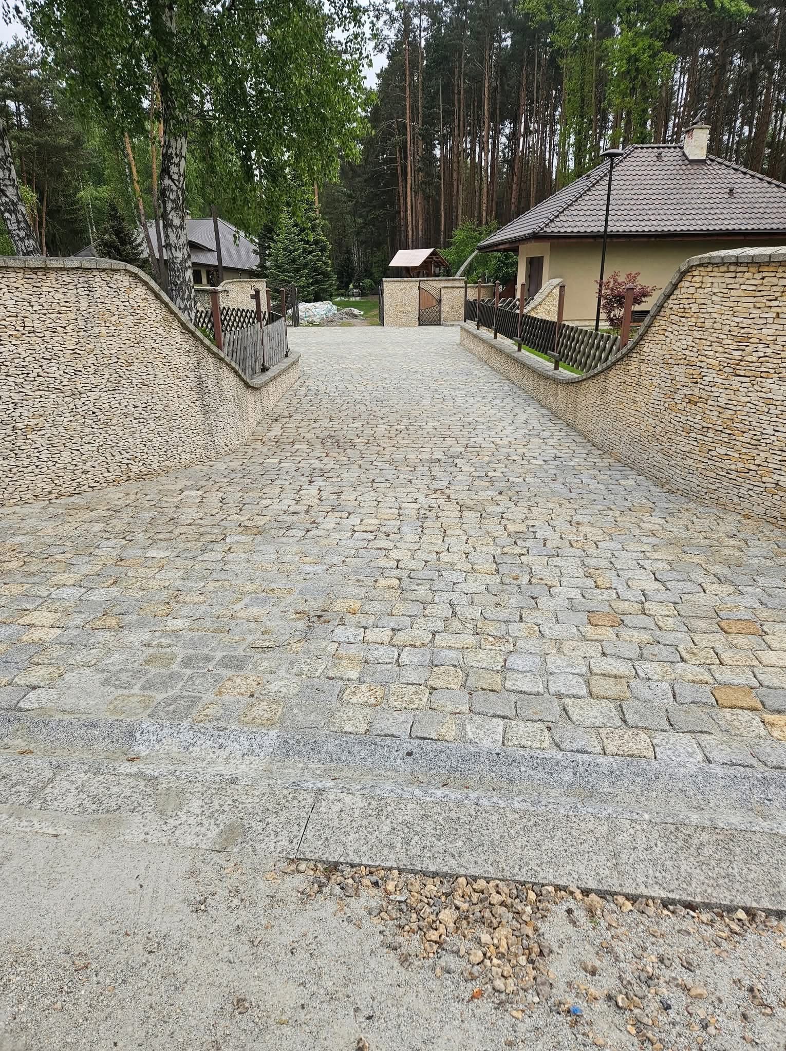 Podjazd z kostki granitowej w odcieniach szarości i beżu, otoczony kamiennymi murkami i zielenią drzew. Widok z góry na posesję.