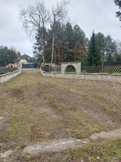 Przygotowany teren pod budowę drogi dojazdowej z elementami ogrodzenia z kamienia i drewnianej siatki. Widoczny dom w tle, pochmurne niebo.