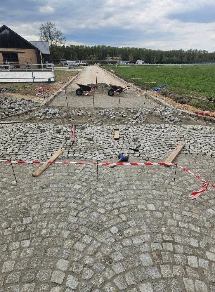 Fragment układanej drogi z kostki granitowej w łuk, zabezpieczonej taśmą. W tle dwie taczki i budynek w otoczeniu zieleni pod pochmurnym niebem.