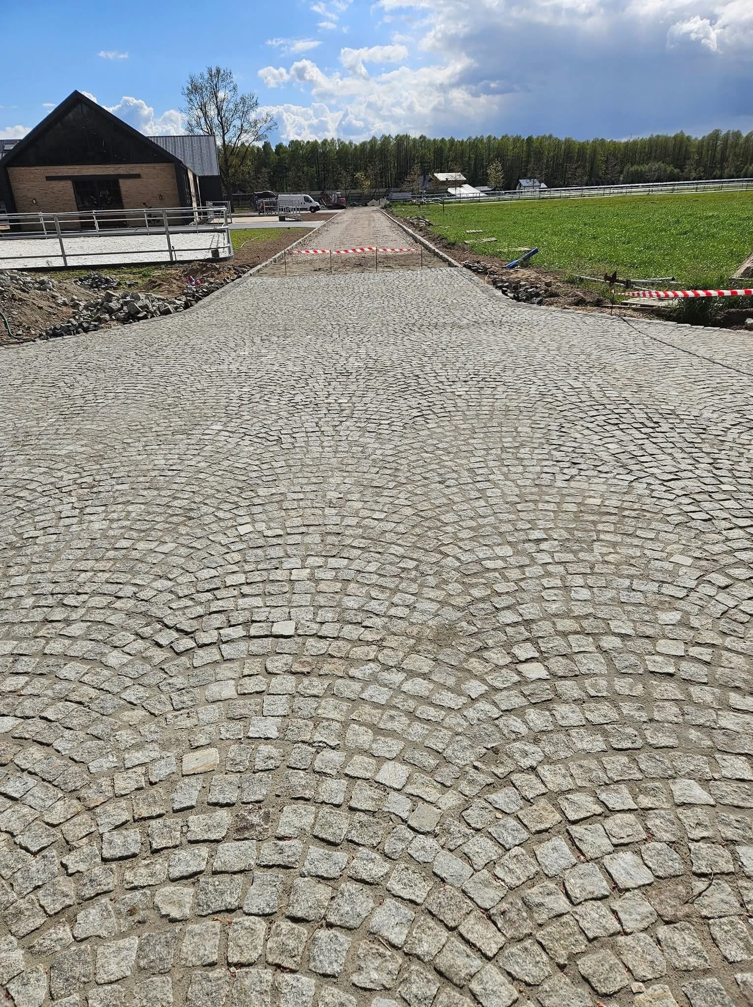 Ułożona kostka granitowa w łukowaty wzór tworzy drogę w kierunku budynku i zielonego pola, pod błękitnym niebem z chmurami.