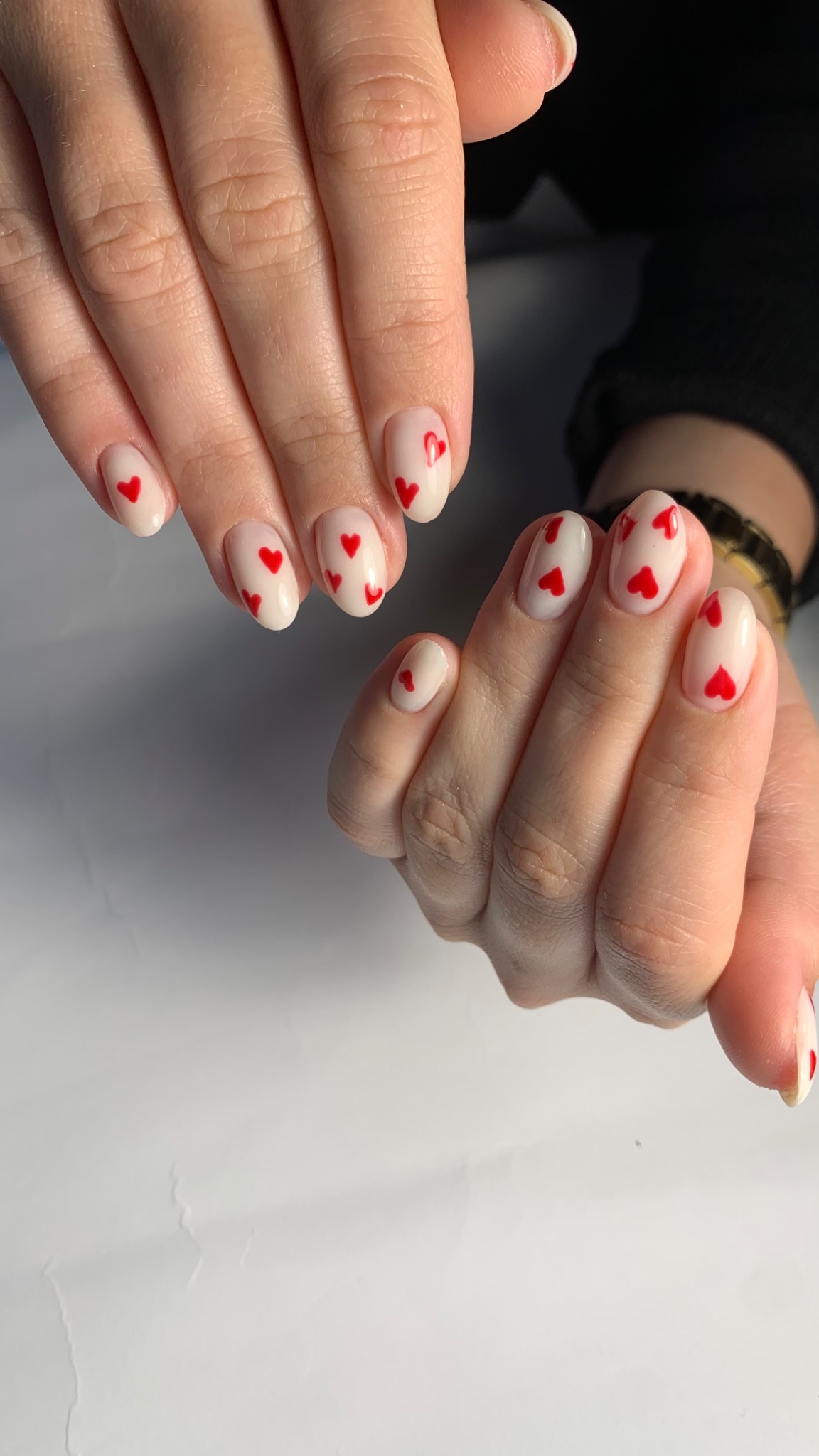 Dłonie z owalnymi paznokciami pomalowanymi jasnym lakierem z drobnymi czerwonymi serduszkami, widoczne na białym tle.
